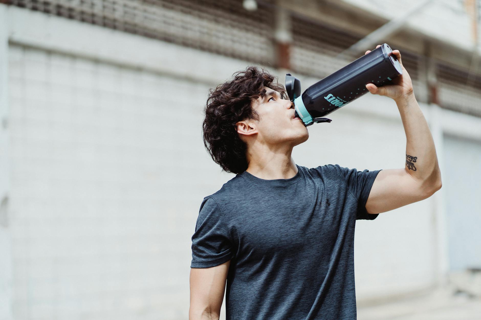 A young man in a casual shirt rehydrates with water from a bottle outdoors. - hydration hacks