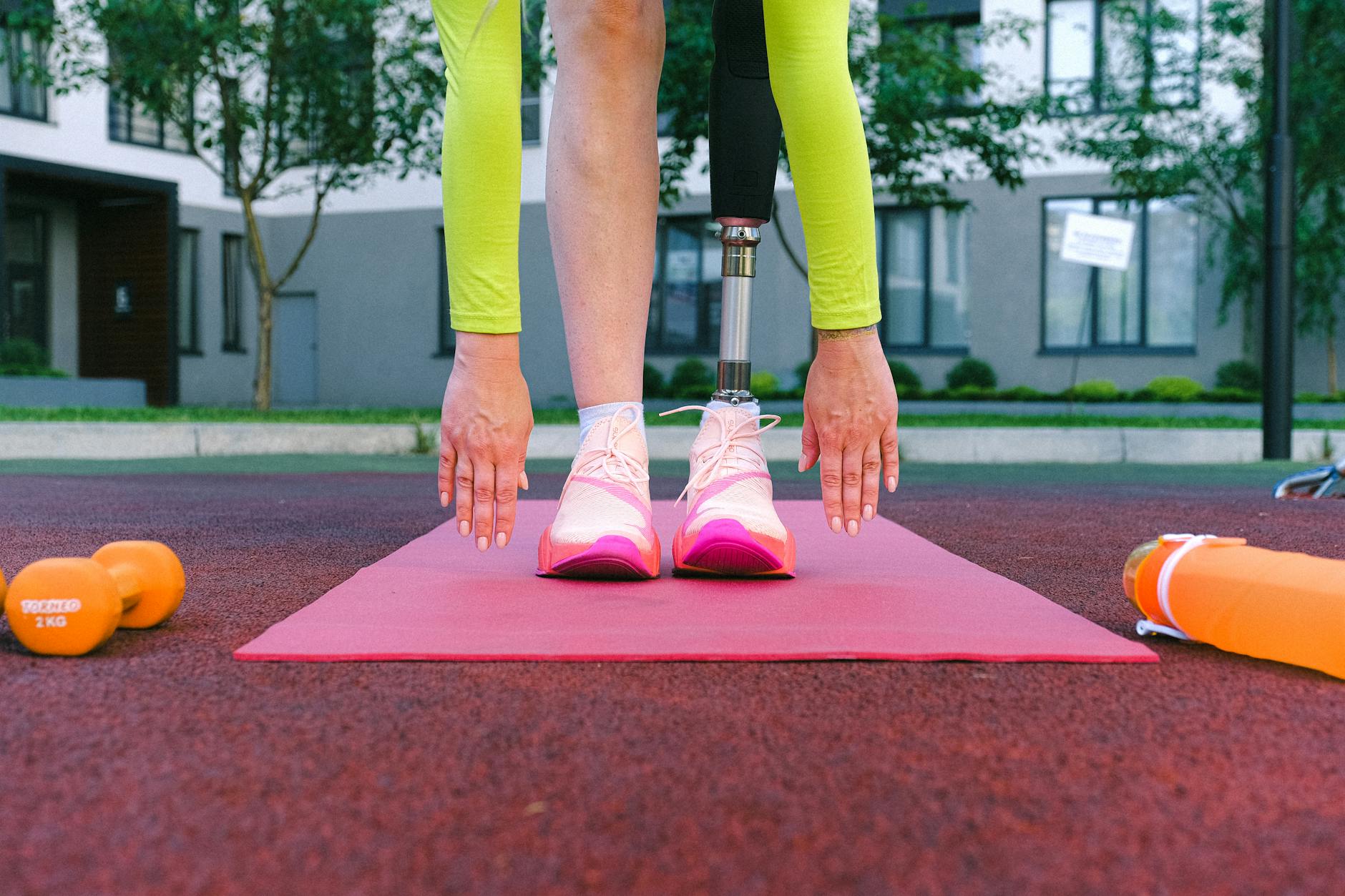 An adult stretching with a prosthetic leg on a red exercise mat outdoors. - lower body exercises