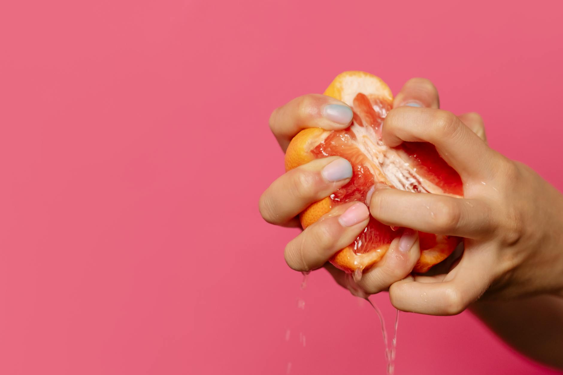 Close-up of hands squeezing juicy grapefruit against a bright pink backdrop, emphasizing freshness. - natural energy boosters