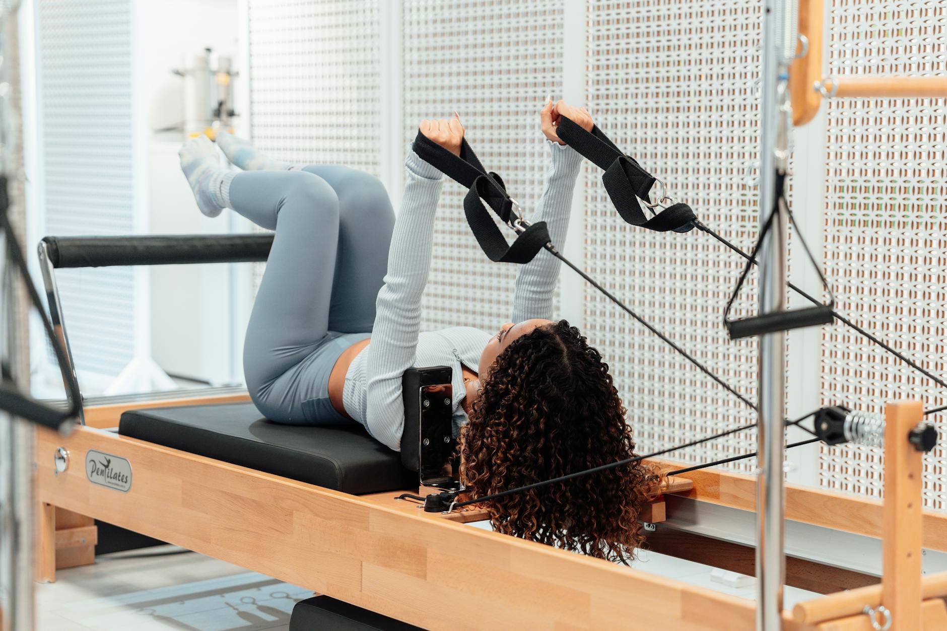 A woman wearing a gray tracksuit exercises on a Pilates reformer in a modern studio setting. - what is pilates
