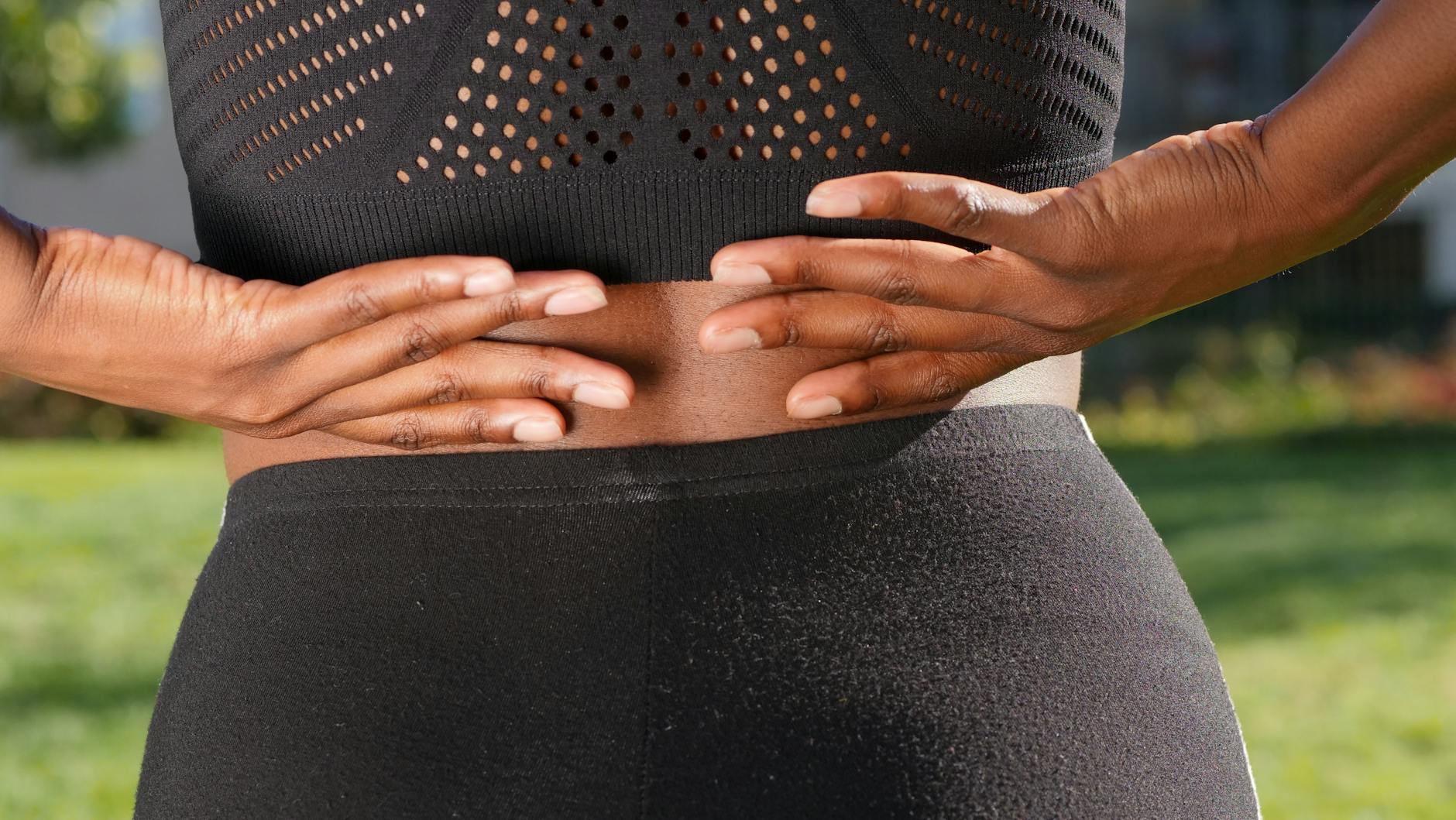 Close-up of a woman's hands gripping her painful back while outdoors. - pregnant back pain