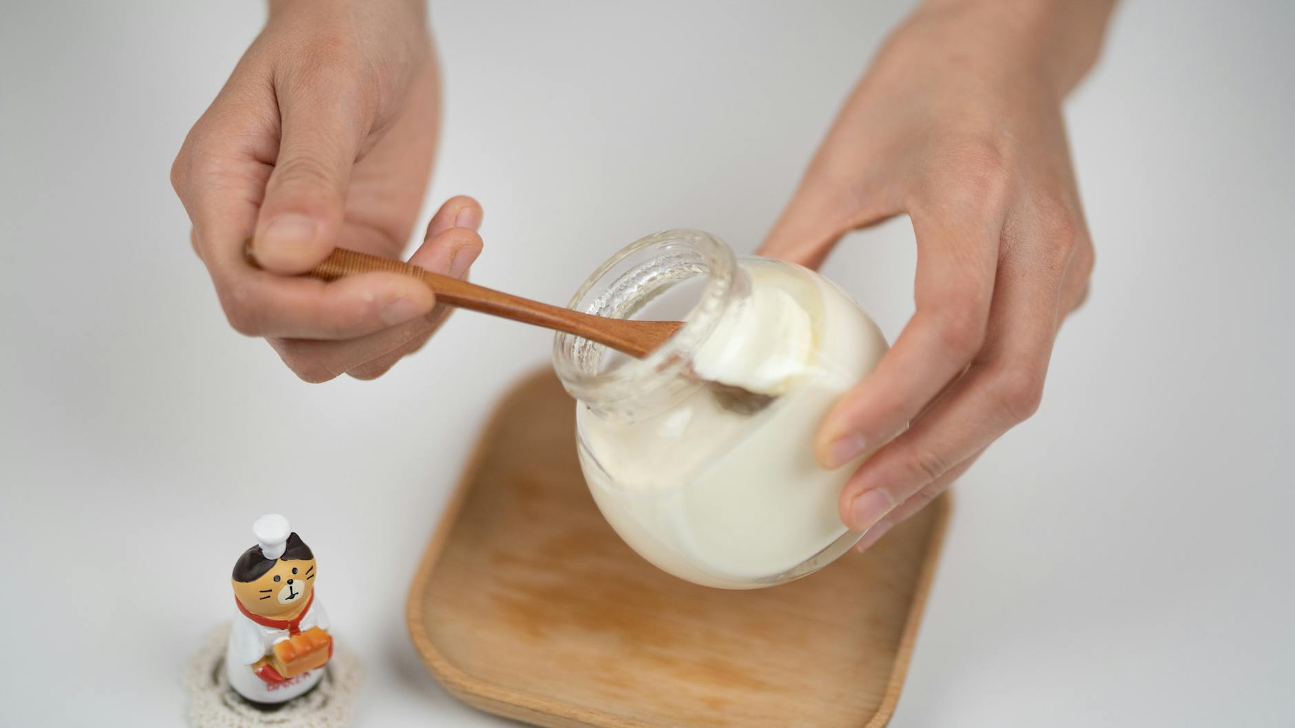 Close-up of hands serving creamy yogurt from a jar using a wooden spoon, with a cute figurine on the side. - probiotics benefits