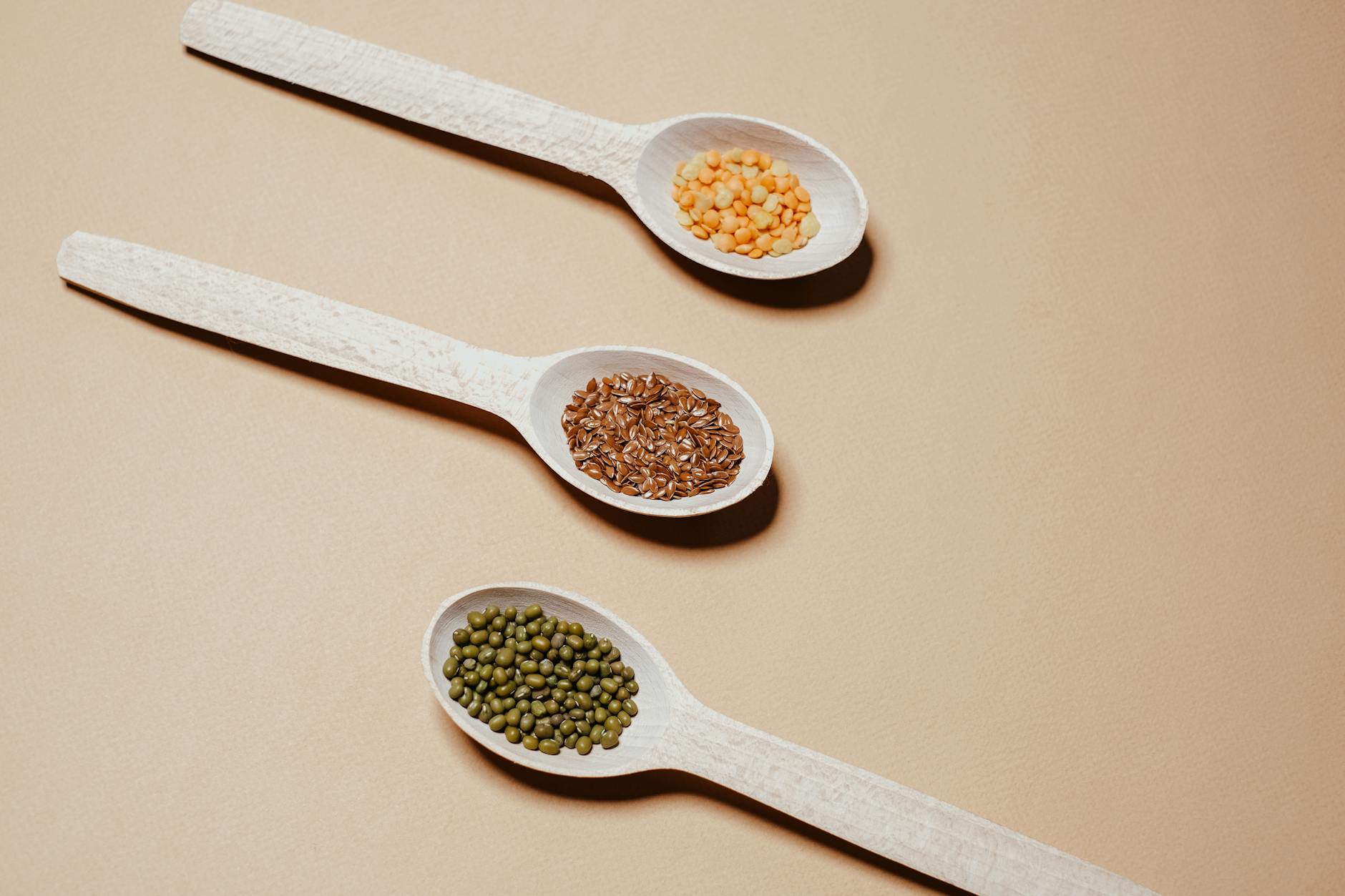 Flat lay of mung beans, flaxseeds, and split peas in wooden spoons, showcasing natural grains. - protein fiber foods