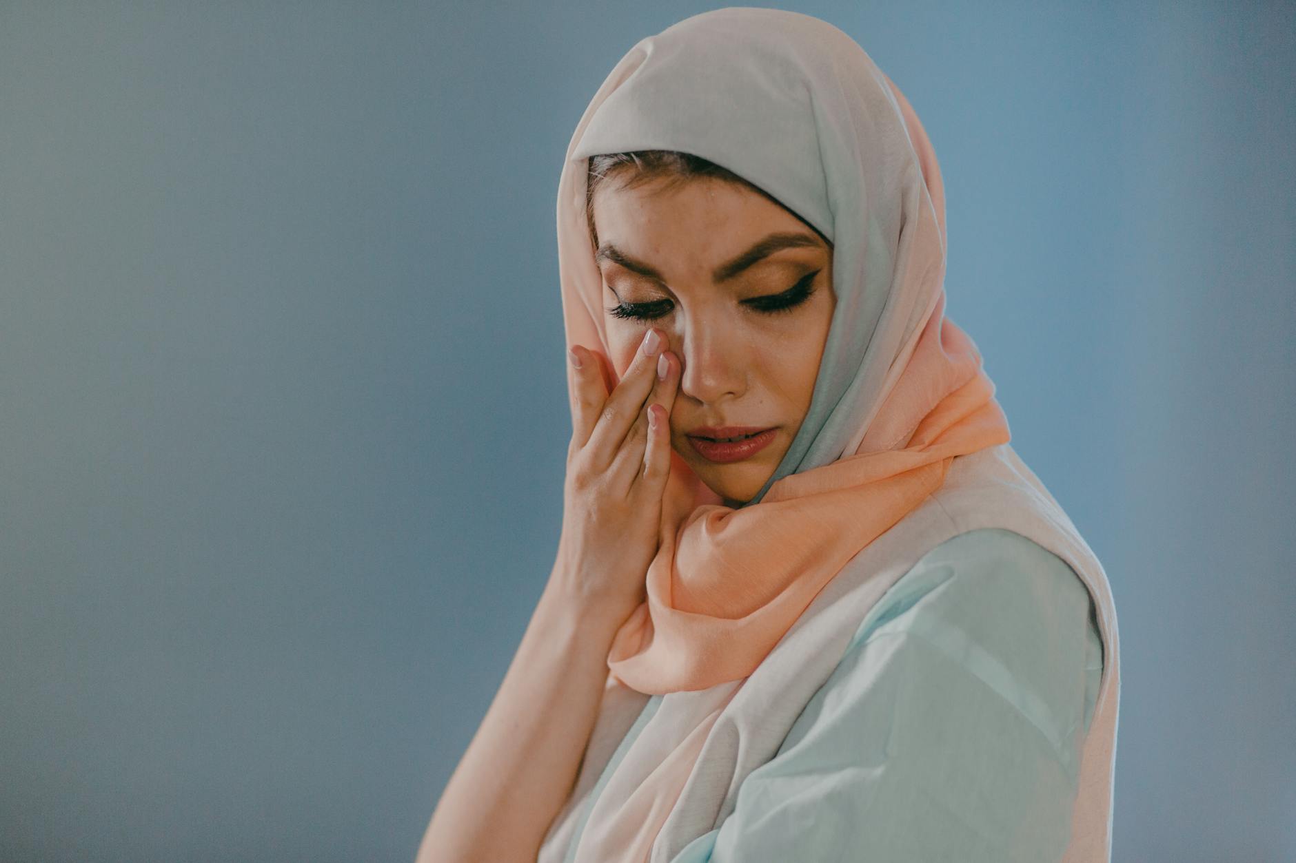 A woman in a hijab wipes tears from her eyes against a blue background. - skinny recipes