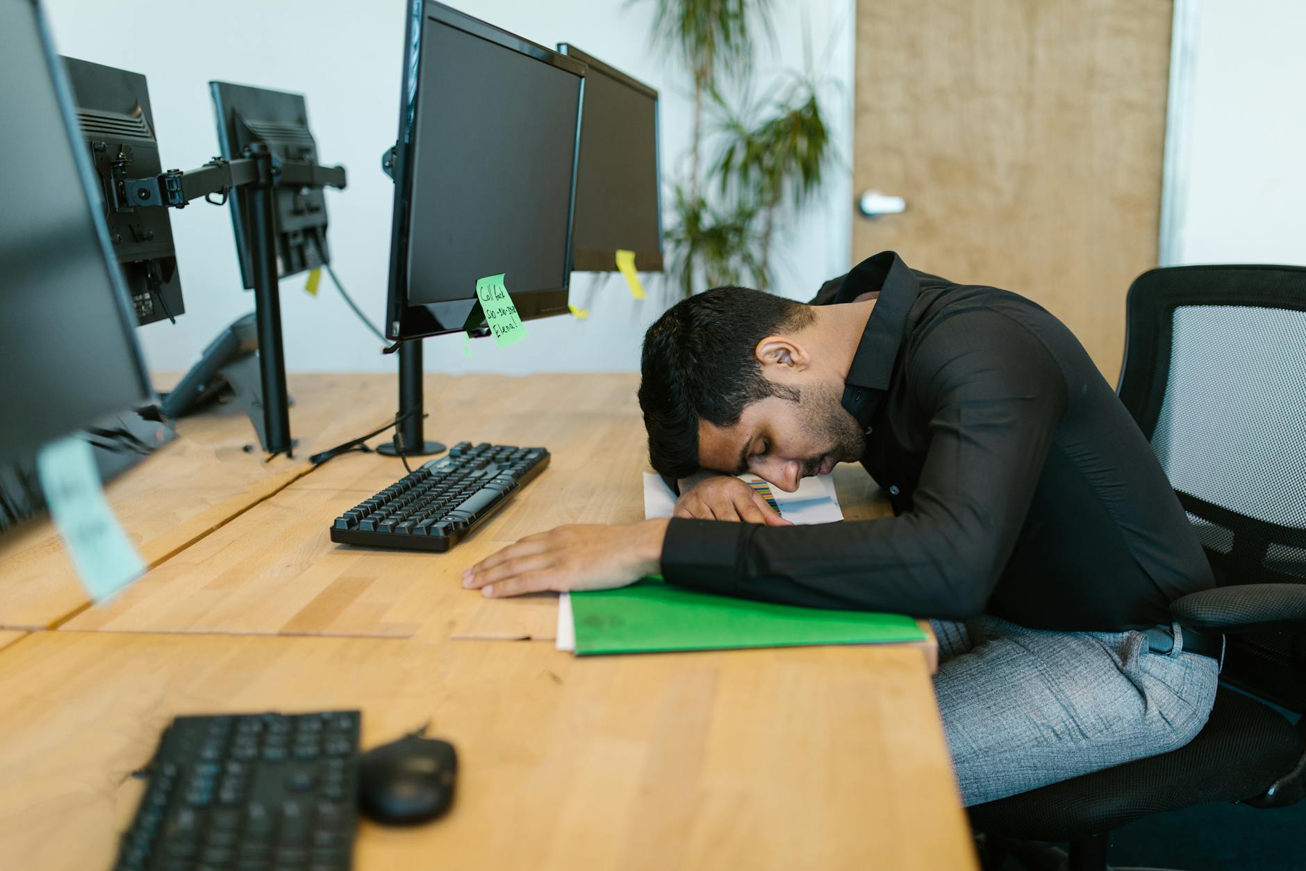 An exhausted office worker resting on a desk surrounded by computer monitors in a modern workspace. - spring fatigue remedies