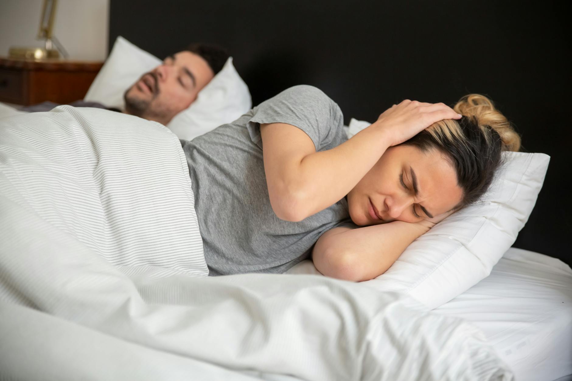 A woman looks frustrated in bed as her partner snores loudly, highlighting sleep disturbance issues. - spring sleep optimization