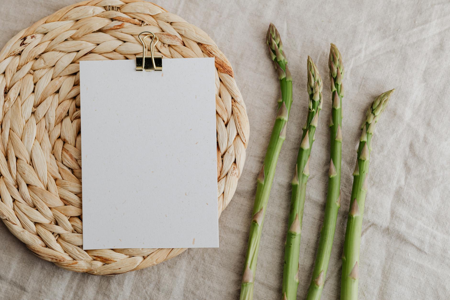 From above of four fresh green asparagus sprouts and blank sheet of paper over round wicker placemat laid on white tablecloth - spring vegetable recipes
