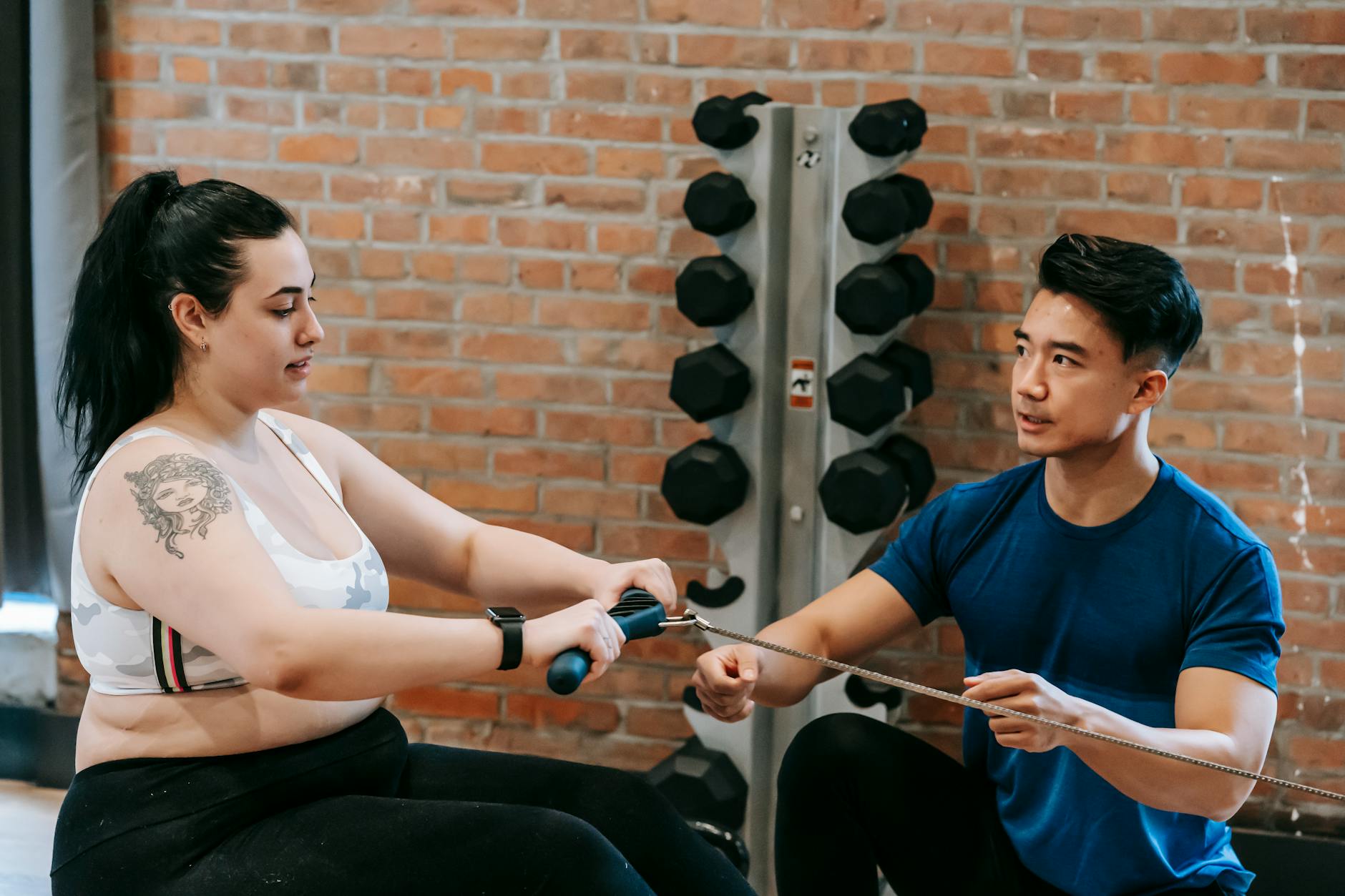Multiethnic trainer and overweight woman doing exercises with rowing machine together in sport club - teen weight loss