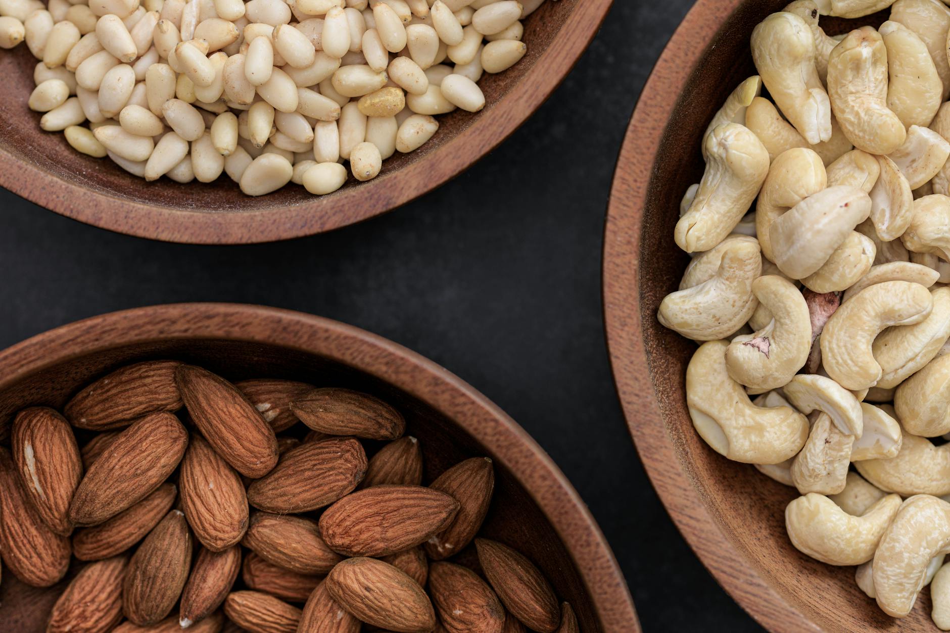 Top view of almonds, cashews, and pine nuts in wooden bowls against a dark backdrop. - vegetarian protein sources
