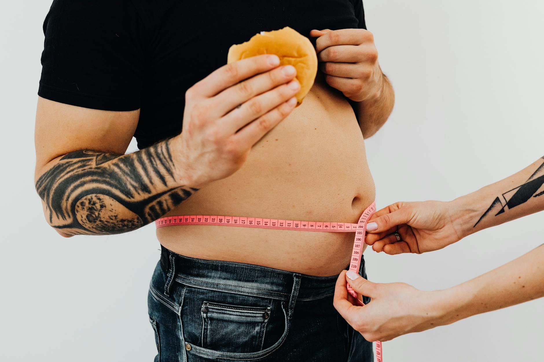 A man with tattoos holding a burger as another person measures his waistline with a tape measure. - weight loss myths