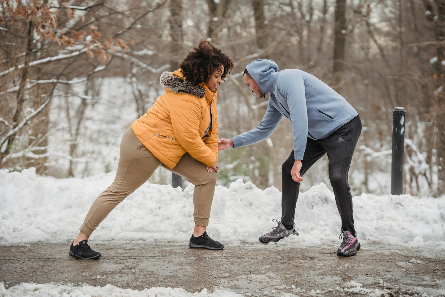 Professional instructor helping African American female doing exercises on frozen path of winter park - winter wellness slump