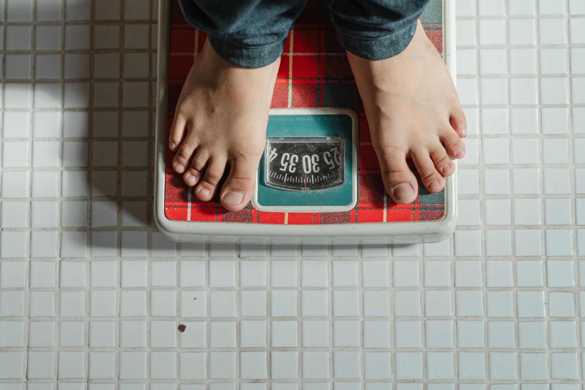 From above crop anonymous barefoot child in jeans standing on weigh scales on tiled floor of bathroom - calorie deficit for weight loss