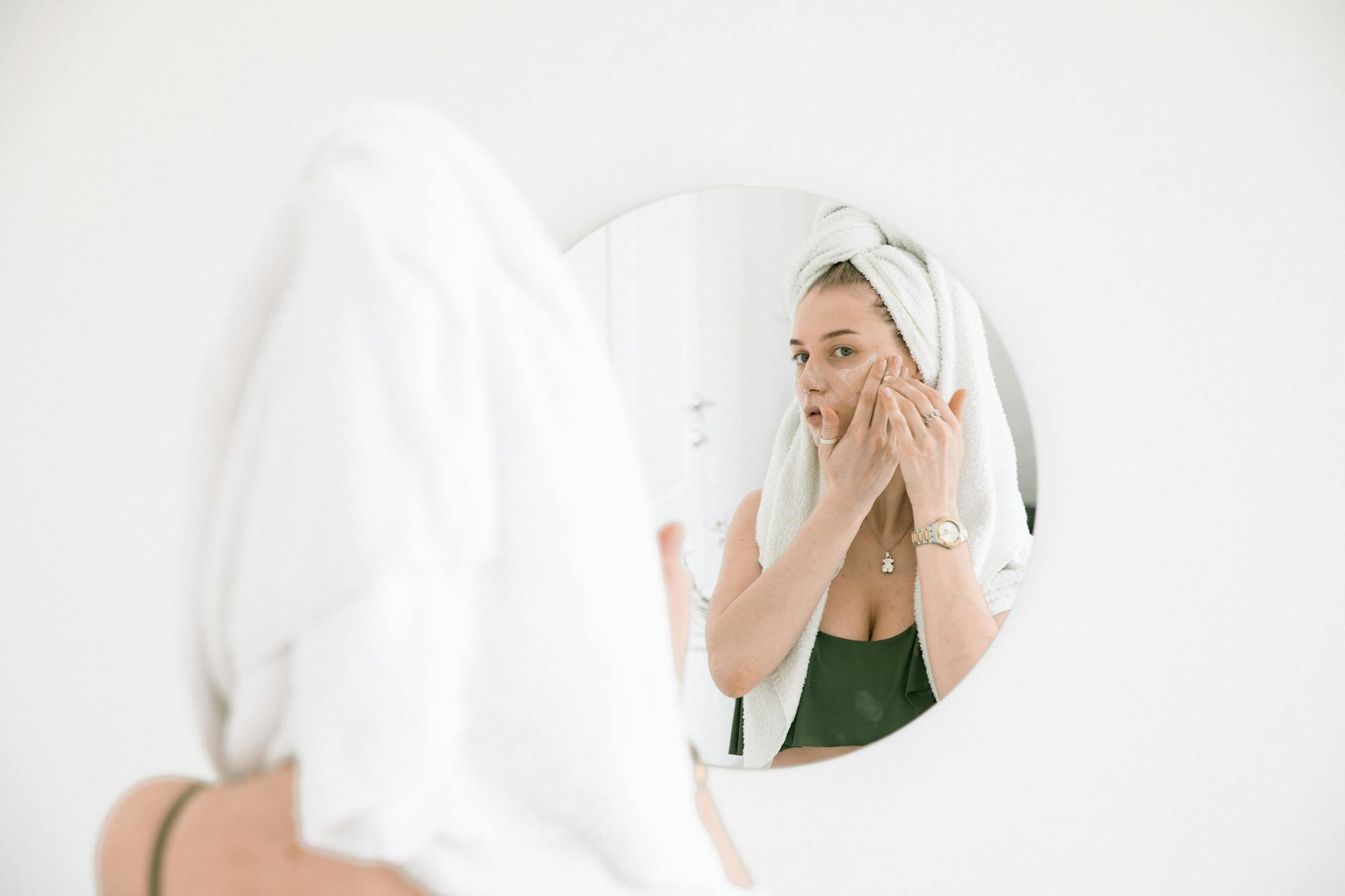 A woman applies face cream in the bathroom, reflecting in a mirror with a towel on her head. - daily skincare routine