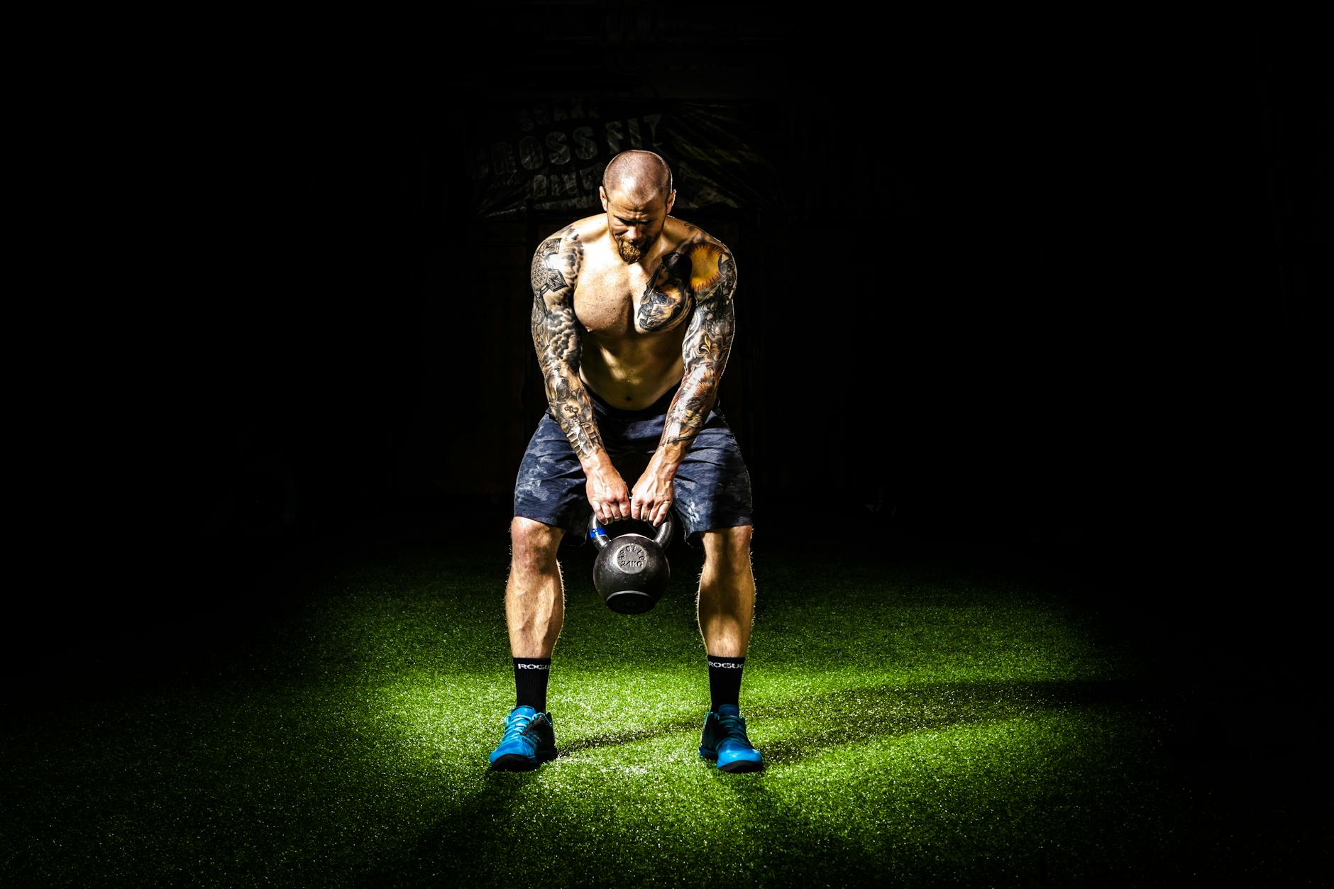 Muscular man with tattoos lifting a kettlebell indoors under a spotlight. Intense fitness and strength training. - gardening strength training