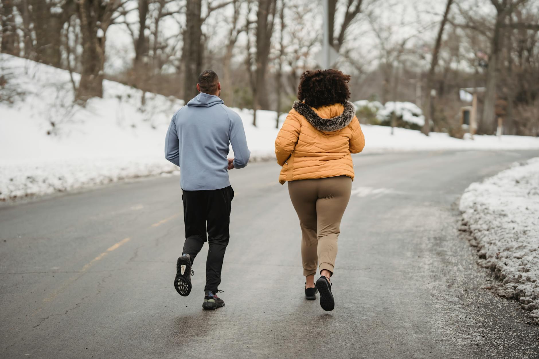 Back view full length fit male fitness trainer and plump female in warm jacket jogging together on asphalt roadway in snowy forested suburb on cold winter weather - post winter workout
