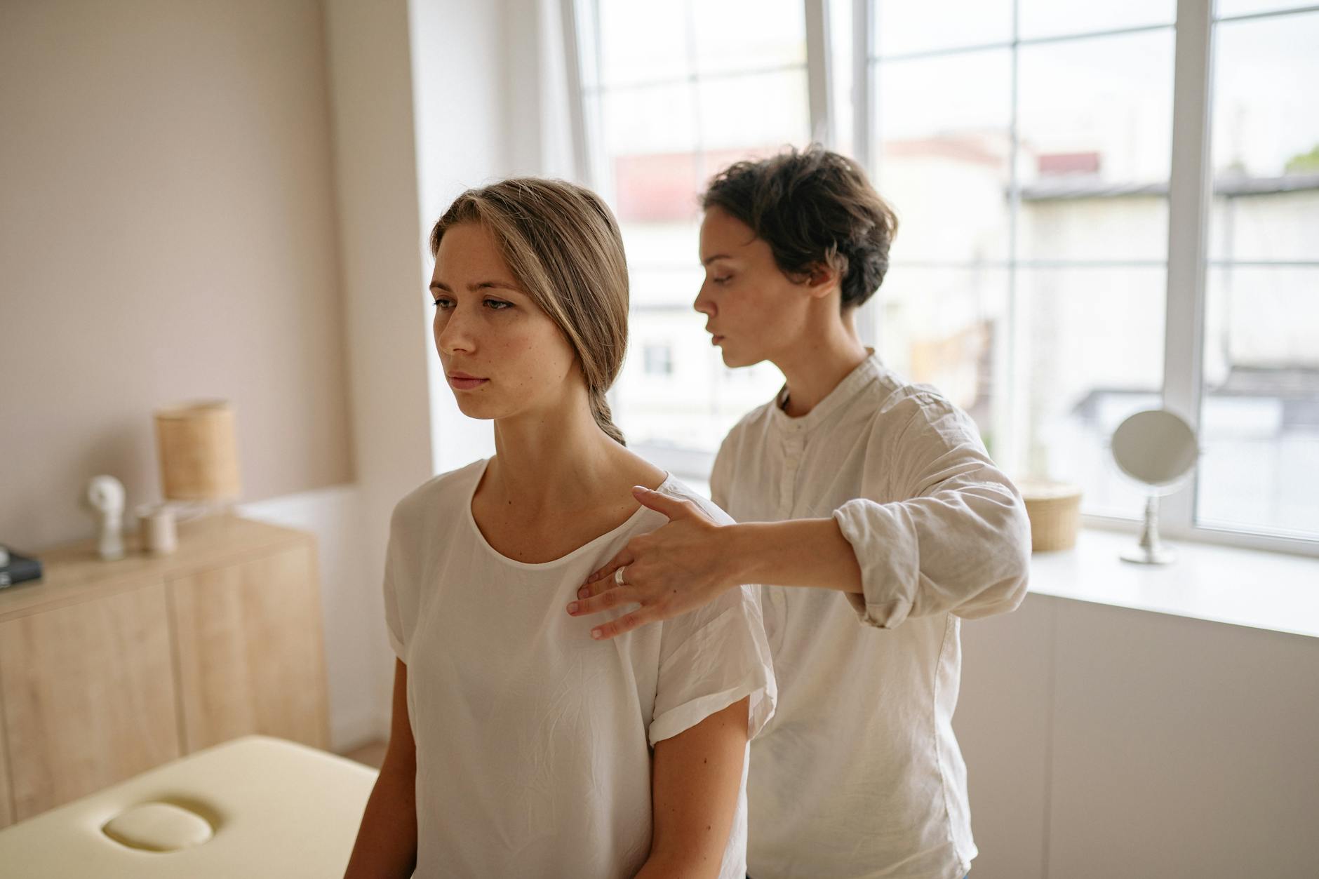 A woman receives shoulder therapy from a therapist in a serene room with natural light. - shoulder pain relief