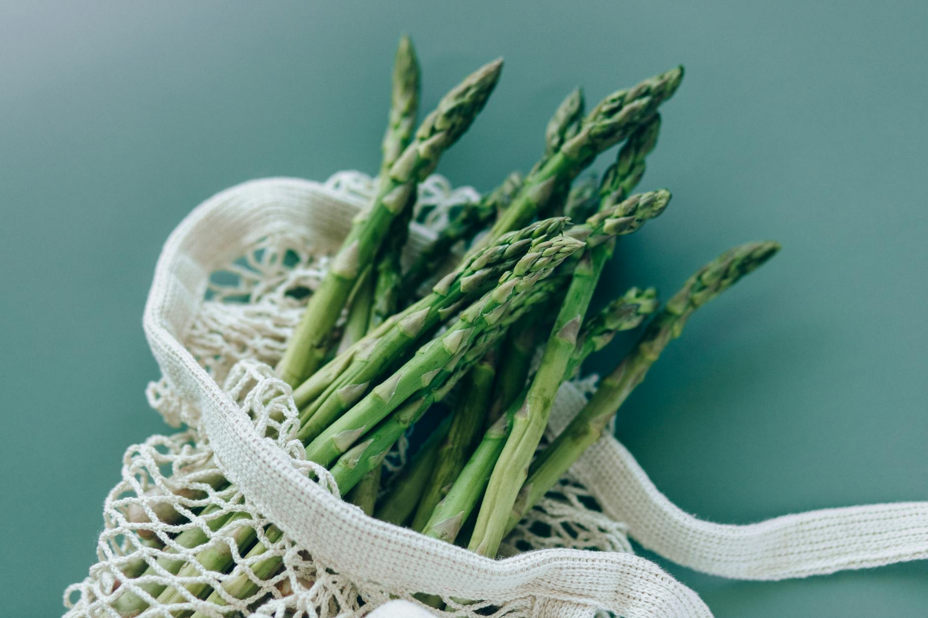 Fresh green asparagus in a reusable mesh bag on a green background, showcasing eco-friendly food storage. - spring energy foods