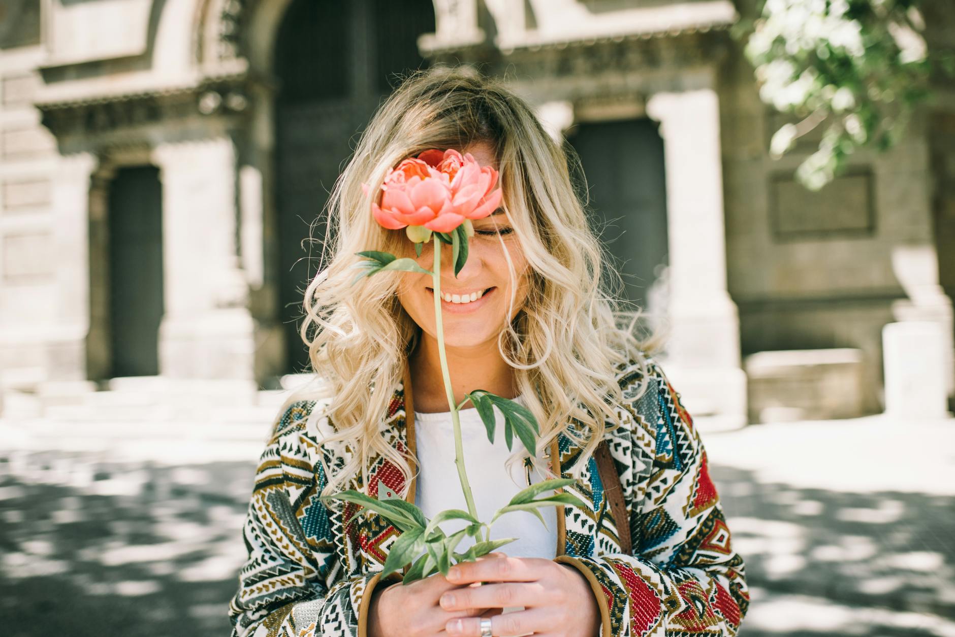 Blonde woman smiling with a flower outdoors, enjoying a sunny day. - spring mood boost