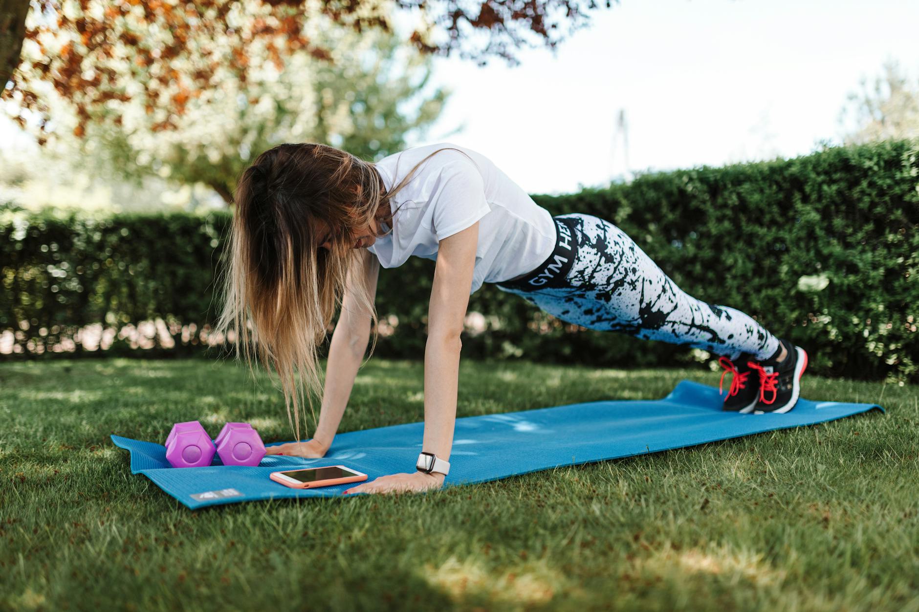 Woman doing push-ups on a yoga mat outside with dumbbells and a smartphone nearby. - spring outdoor workouts