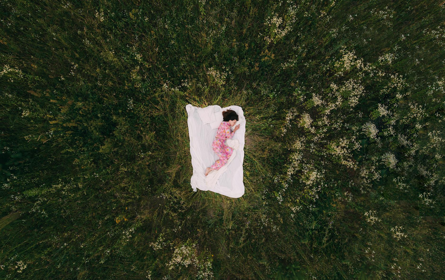 A peaceful aerial view of a person in pajamas sleeping on a blanket in a grassy field. - spring sleep reset