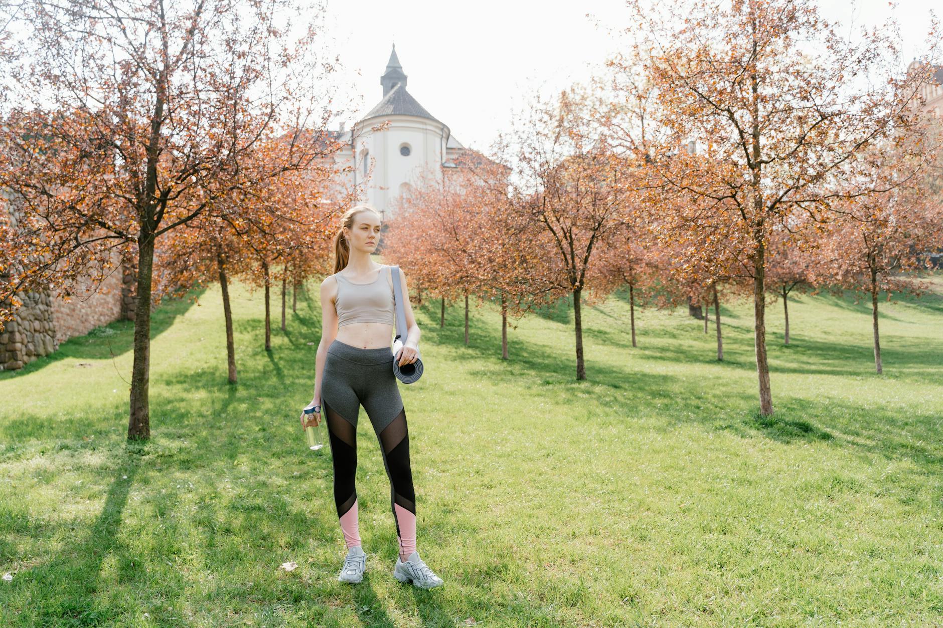 A woman stands in a sunny park, holding a yoga mat and water bottle, surrounded by trees. - spring wellness tips