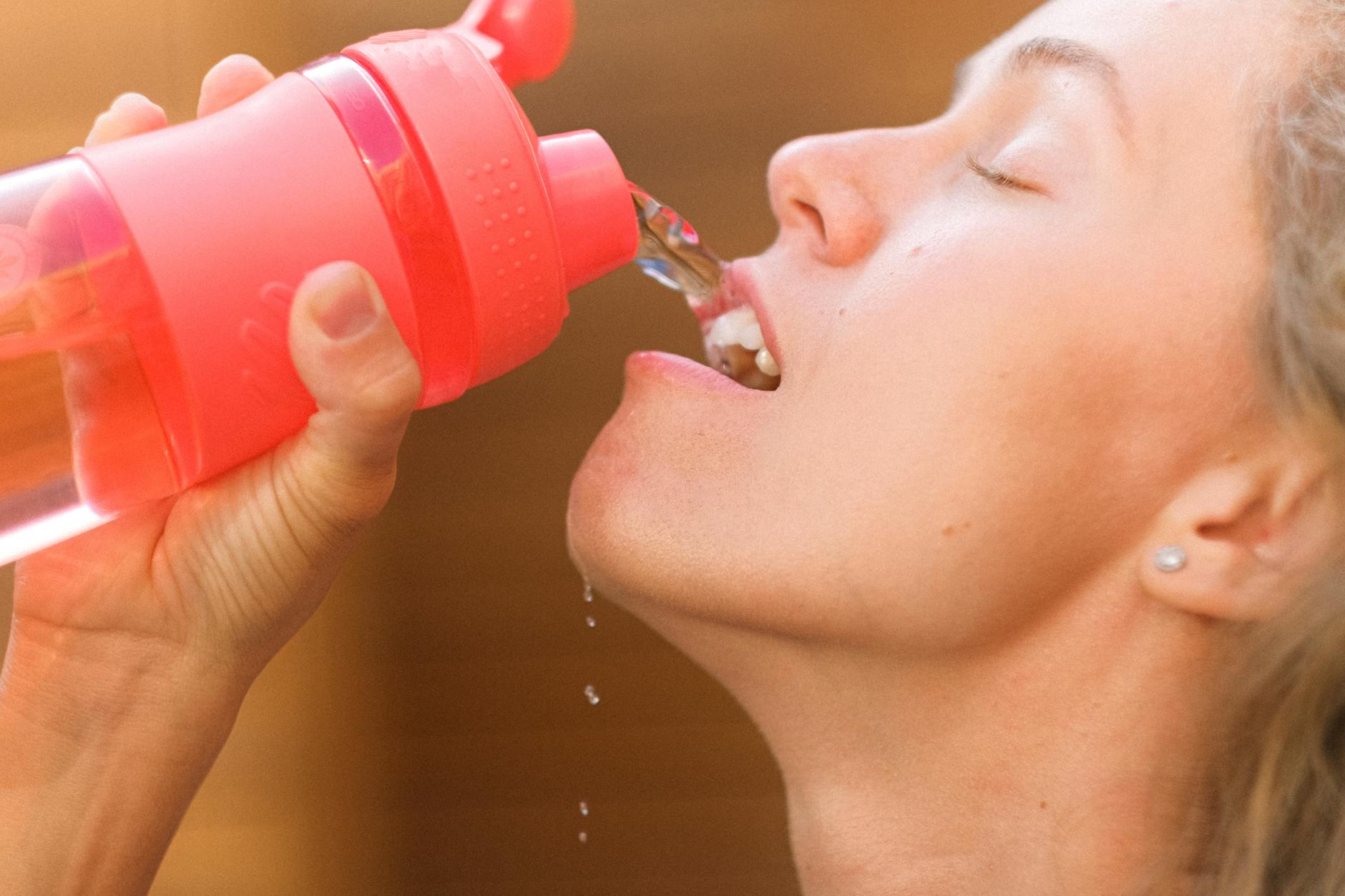 Crop tired young female pouring refreshing clean water into mouth from reusable bottle after intense training - staying hydrated spring