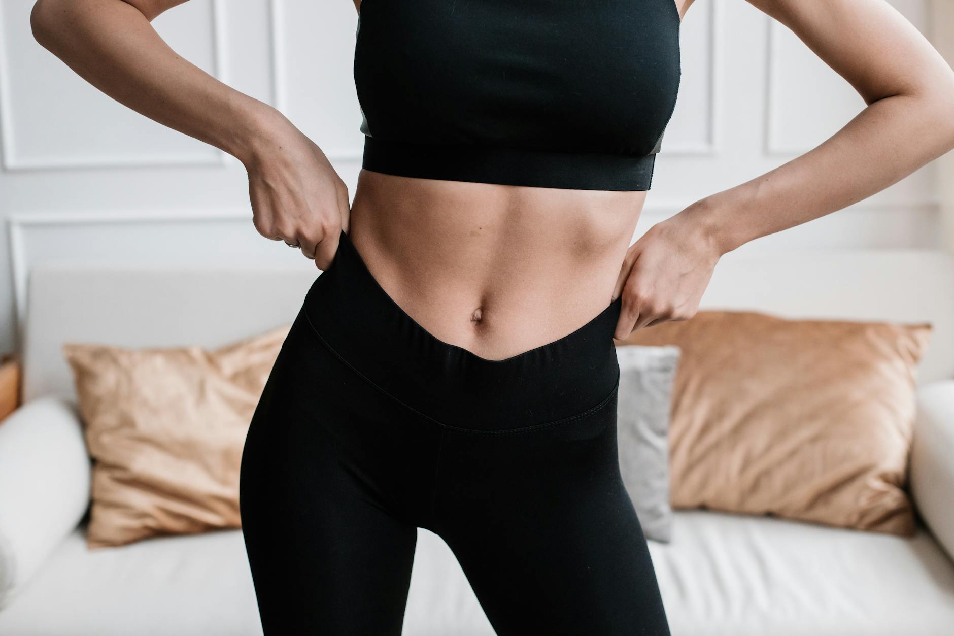 Close-up of a woman in activewear adjusting her pants in a stylish living room. - stomach fat workout