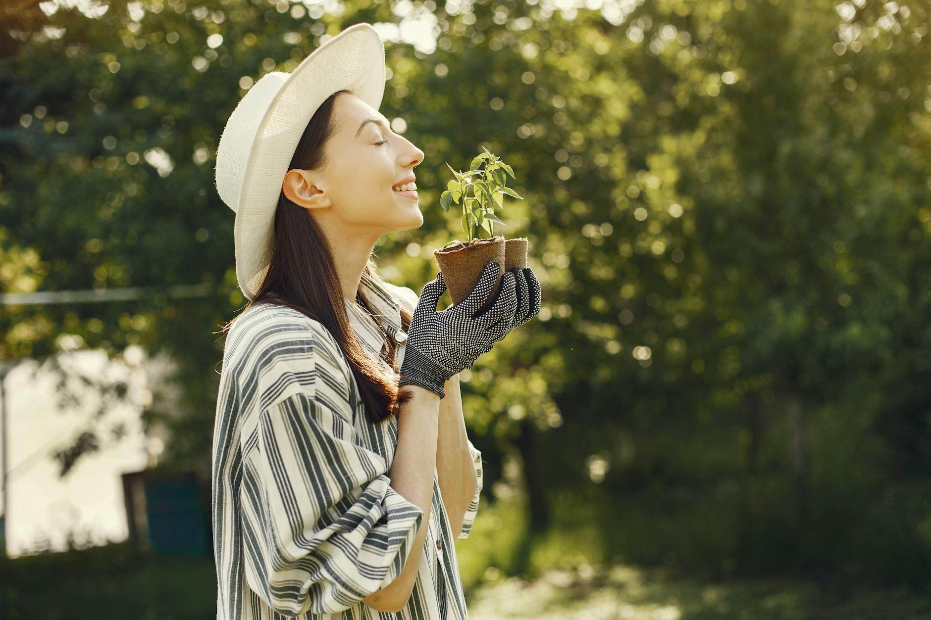 Smiling woman in striped shirt and hat holding a potted plant in a sunlit garden. - gardening for stress relief