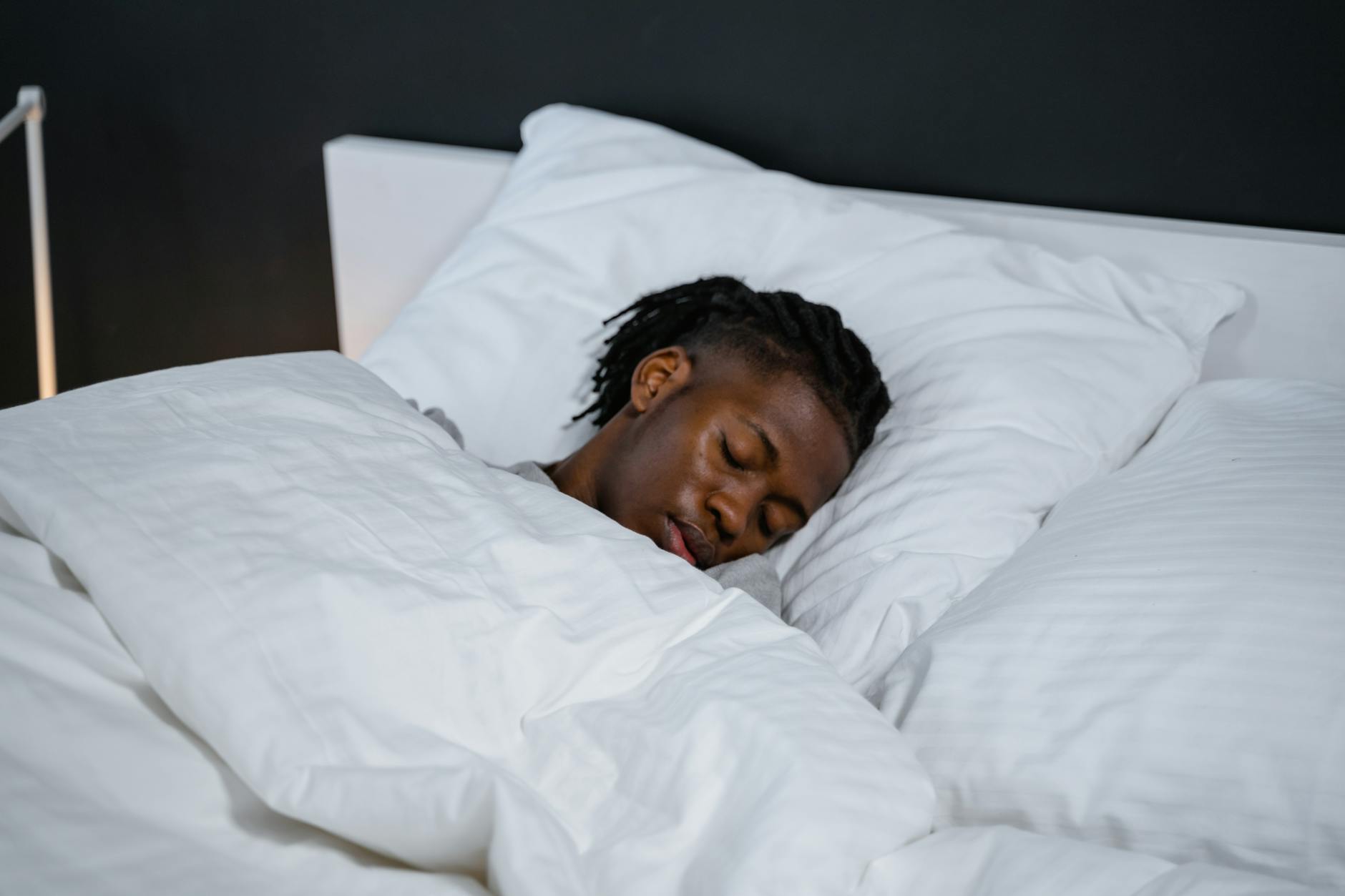 A young man resting peacefully in a bed with white sheets and a dark background. - magnesium for sleep
