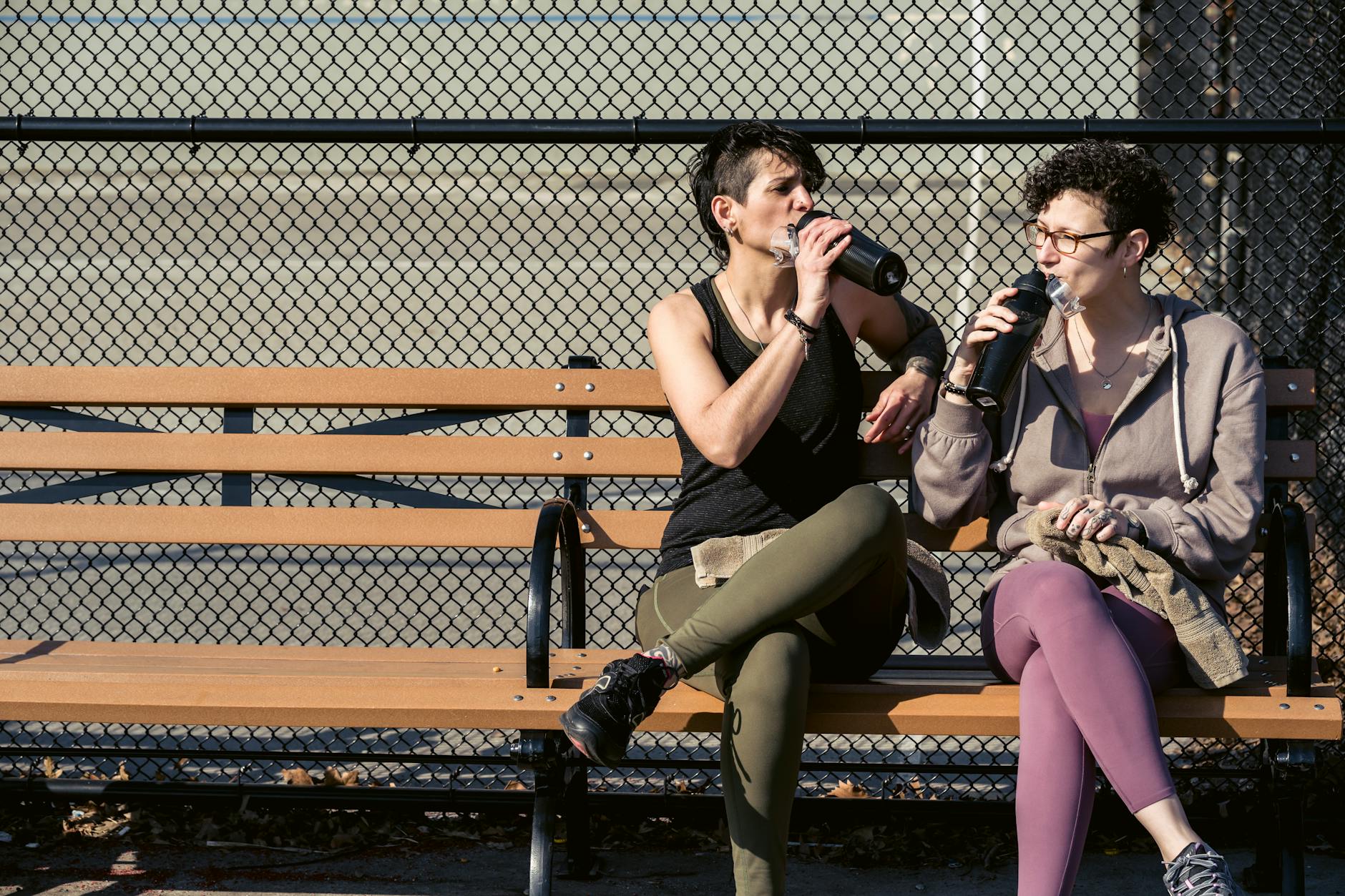 Active female friends in sportswear sitting on bench and drinking water from bottles while resting after workout in park - outdoor workout hydration