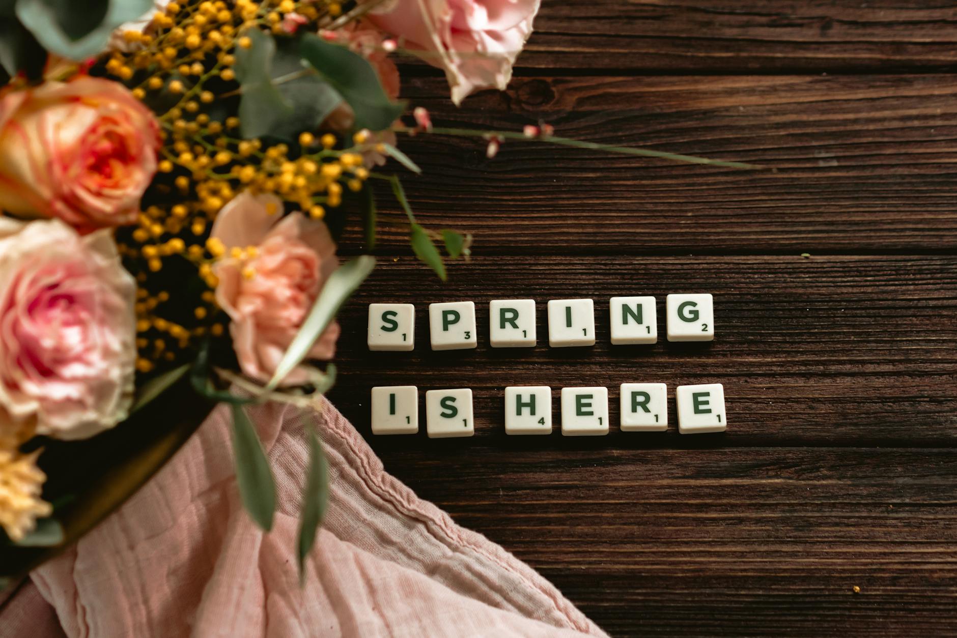 A close-up of roses and spring flowers next to 'Spring is Here' spelled with Scrabble tiles. - spring cravings explained
