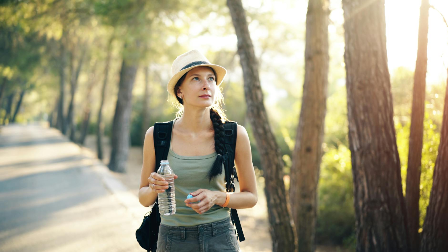 Woman with braid and hat hiking in a forest, holding a water bottle during a sunlit evening. - spring hike hydration