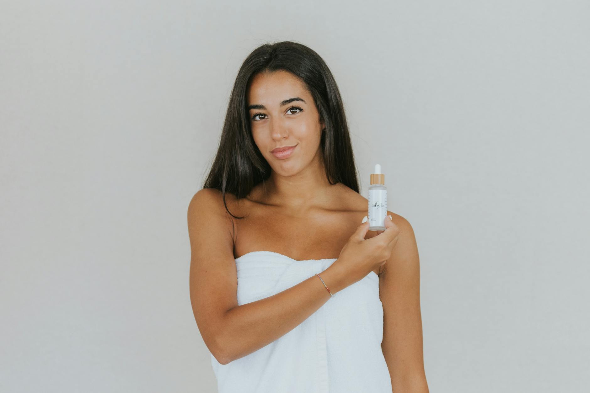 Young woman with long hair holding a skincare serum bottle against a light grey background. - spring skin glow