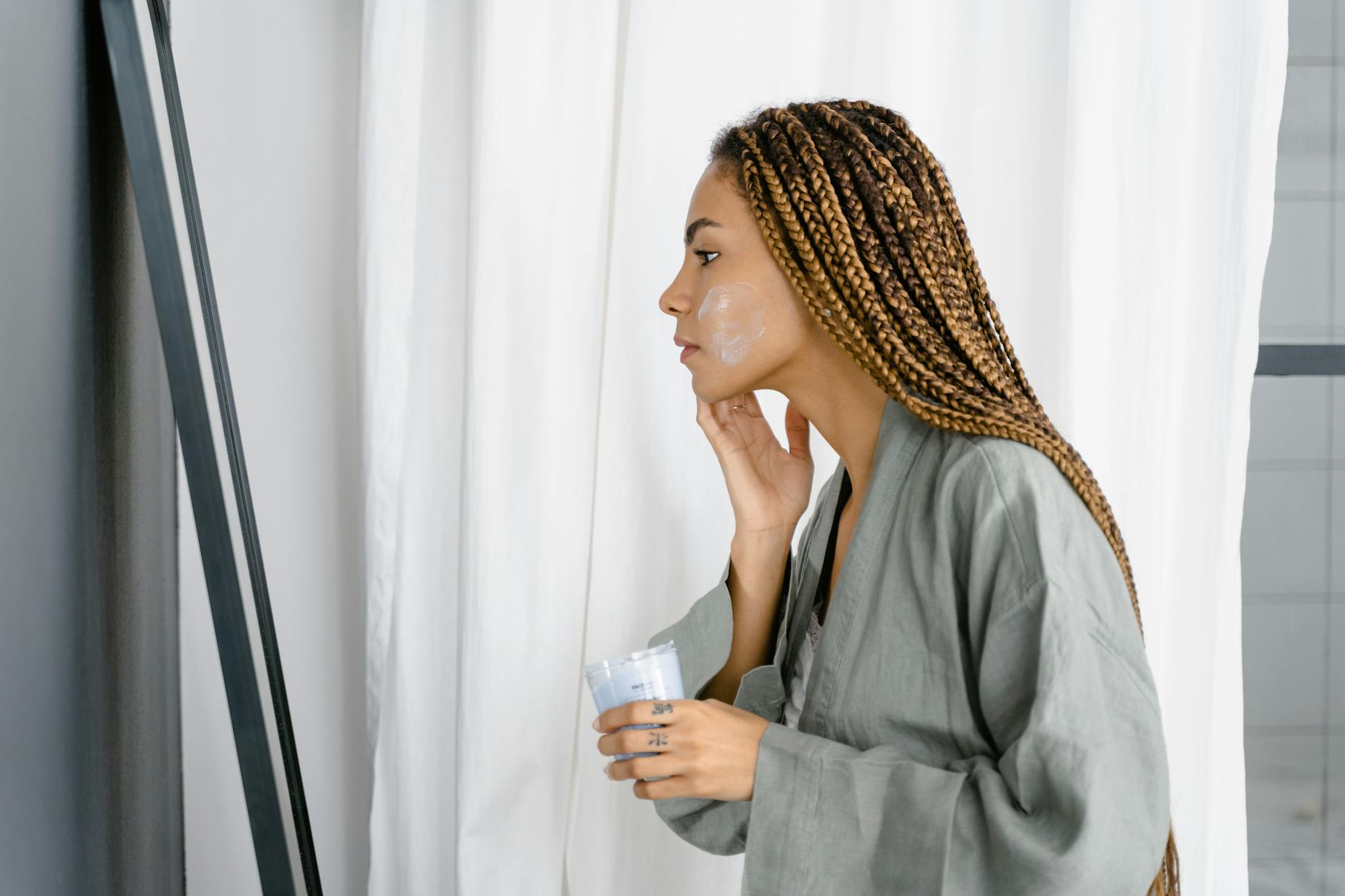 A woman in a green robe applying skincare cream in front of a mirror indoors. - spring skincare tips