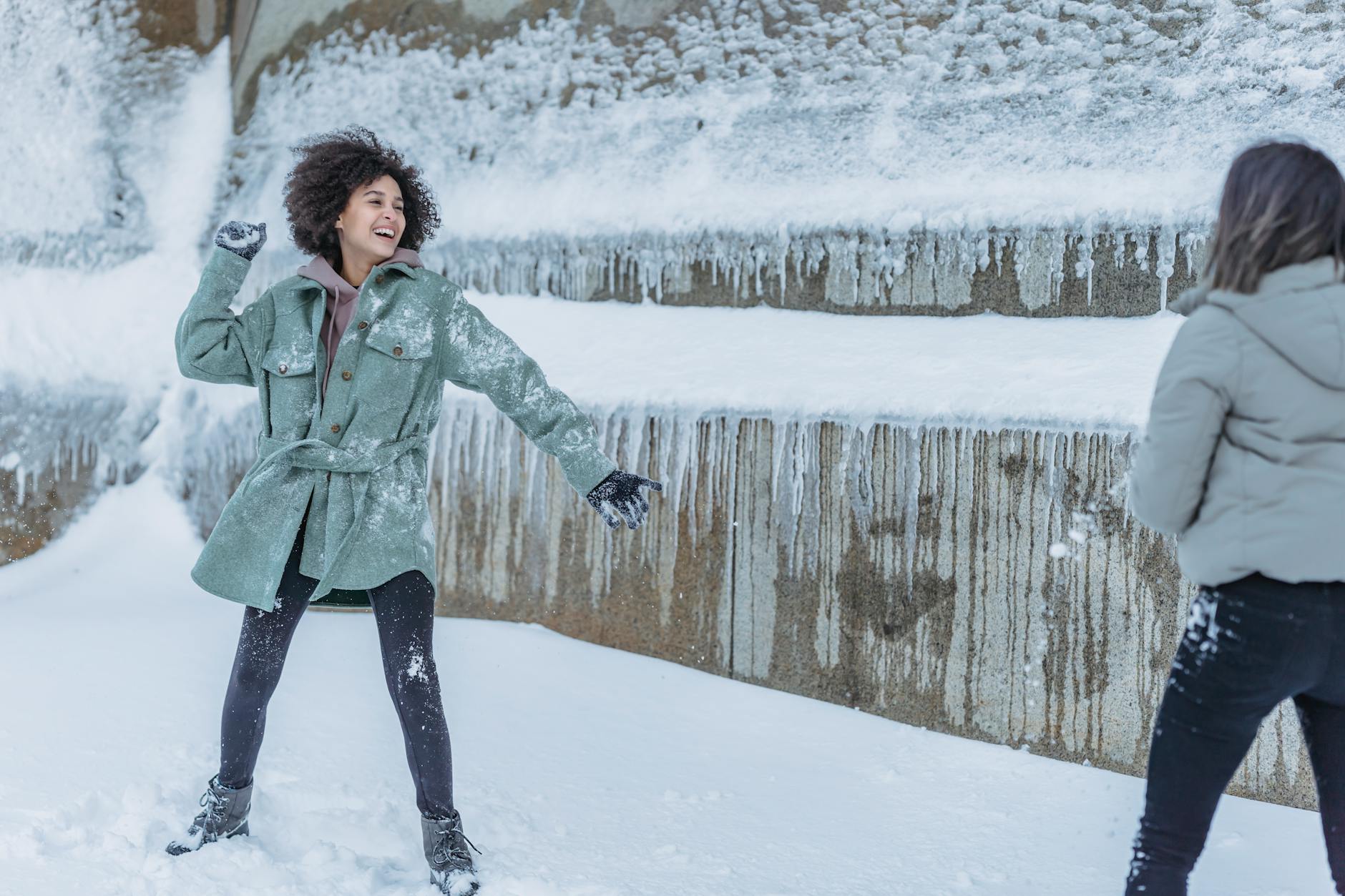 Laughing female friends in warm clothes standing near stone wall covered with snow and playing snowballs in winter - winter body reset